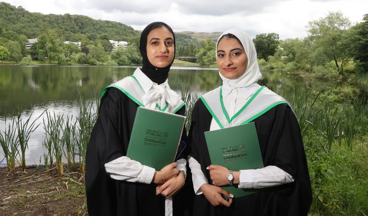 Two students pose in graduation gowns by the loch