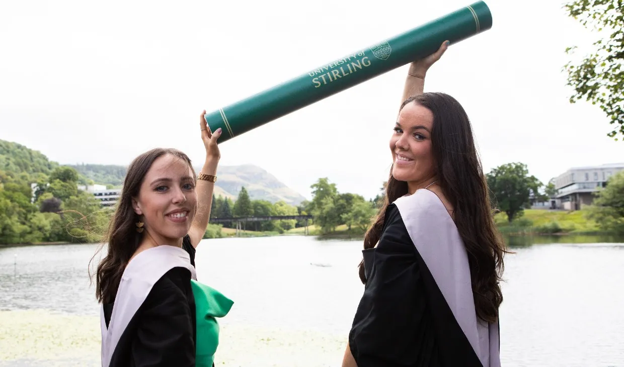 Two female graduates pose by the loch holding a giant scroll prop