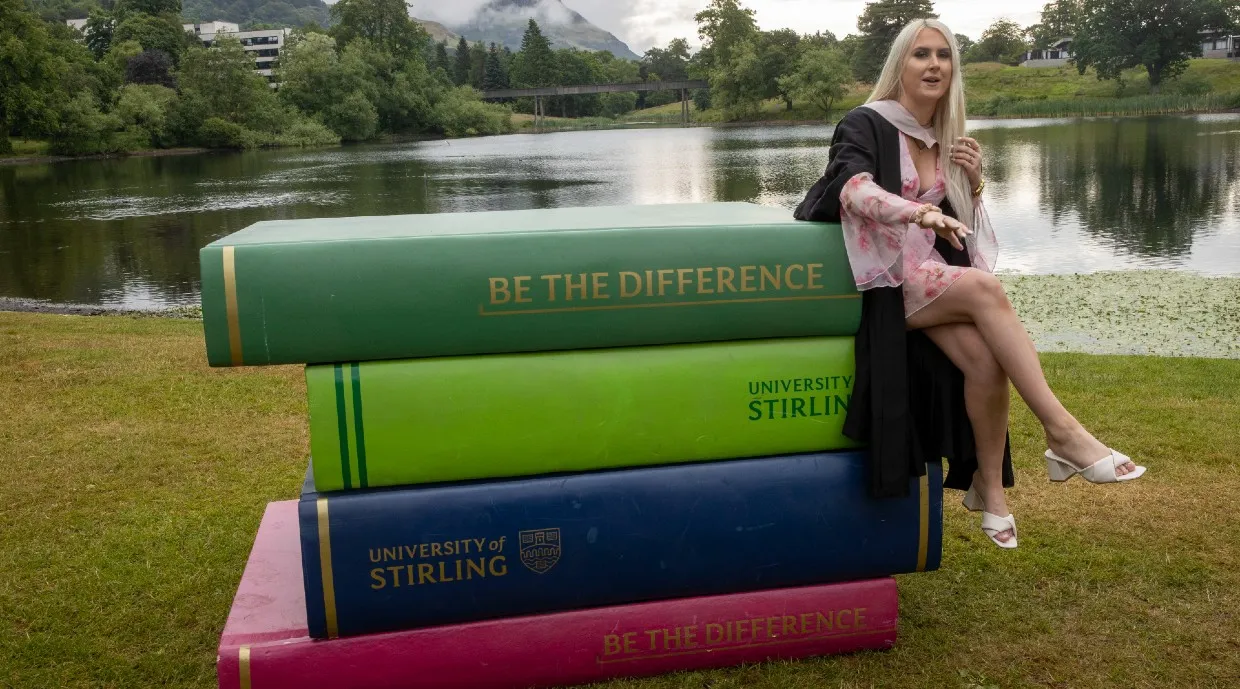 Female graduate perched on large book props by loch