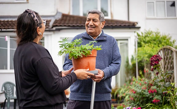 Two older adults talking about a pot plant outside a house