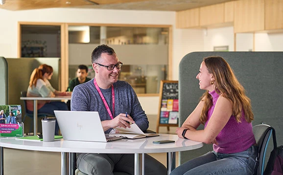 Adult providing student support to a student. They are sitting at a table with a laptop and a notebook.