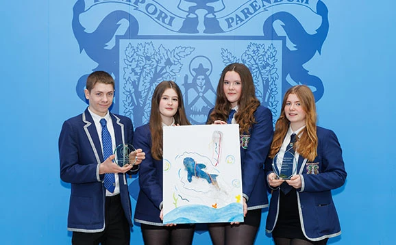 School pupils hold artwork and environmental award with adults. They pose in front of the school crest