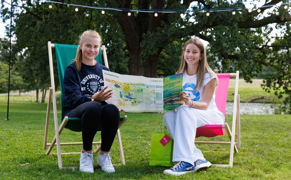 Two smiling students sitting in deckchairs on a grassy lawn, holding a fold-out campus map between them under string lights.
