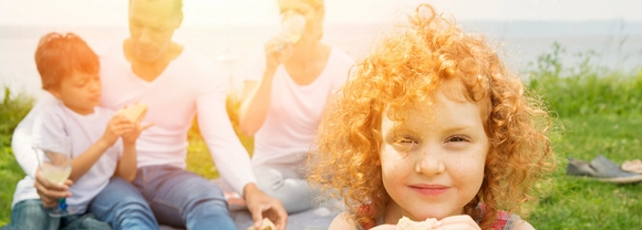 Girl with red, curly hair eating a sandwich with man and woman in the background