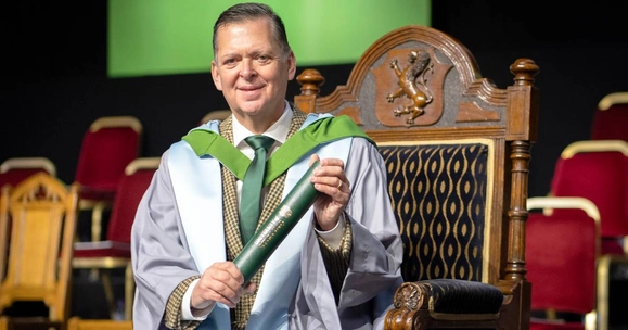 Stanley Morris sits on stage at the University of Stirling graduation ceremony. He wears graduation robes and holds a graduation scroll.