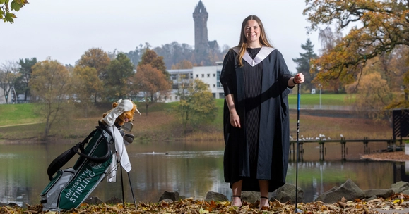 Lorna McClymont posed with golf club in front of University of Stirling loch, with Wallace Monument in background.