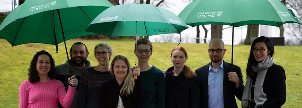 Group of eight people standing under three University umbrellas