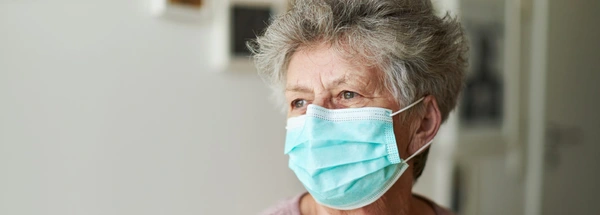 Older person wears a mask and is gazing out of a window