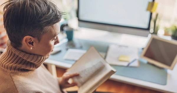 woman reading book at desk in front of computer