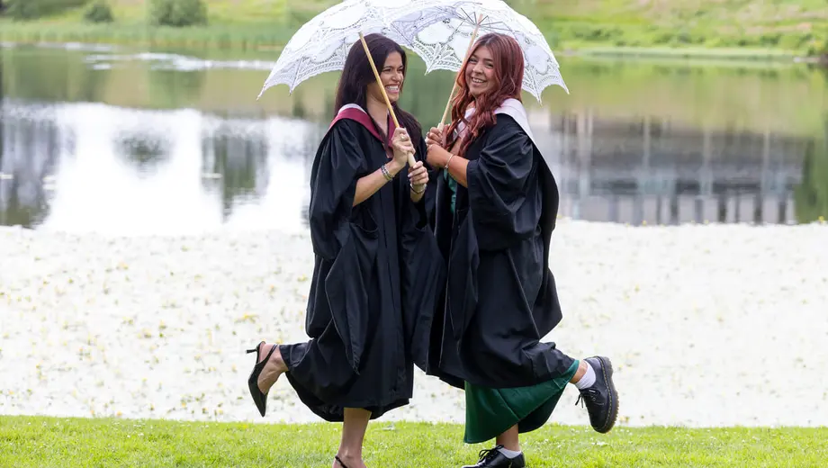 Two students in graduations gowns pose with parasols by loch.